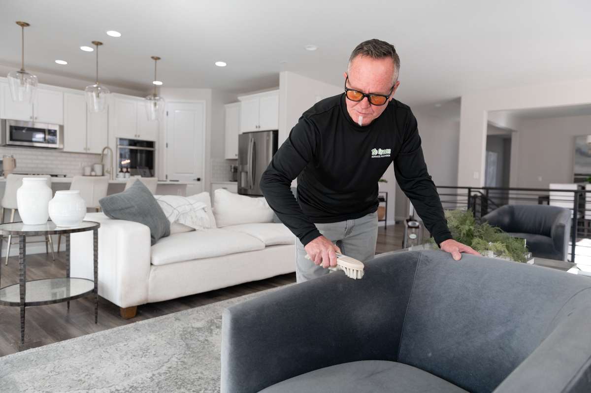 A professional carpet cleaner using a natural fiber brush to dust a grey arm chair during cleaning