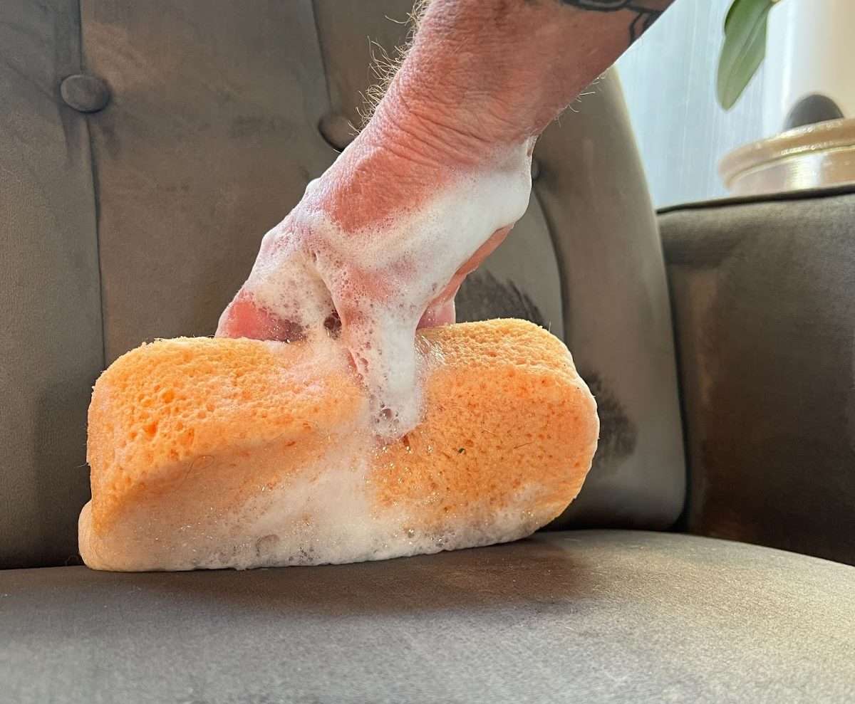 A professional carpet cleaner using natural products to create foam and apply to a grey couch with a sponge.